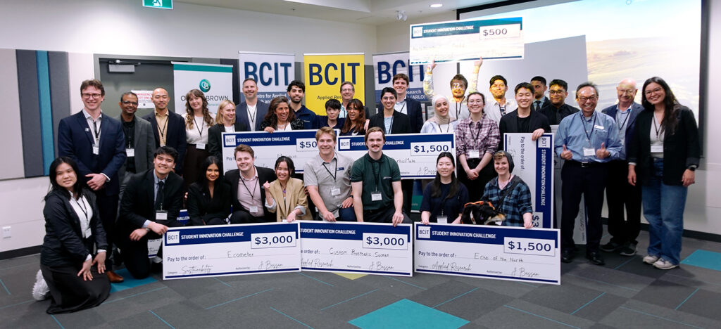 A group of over 30 people grouped in fron of three BCIT banners holding large novelty cheques