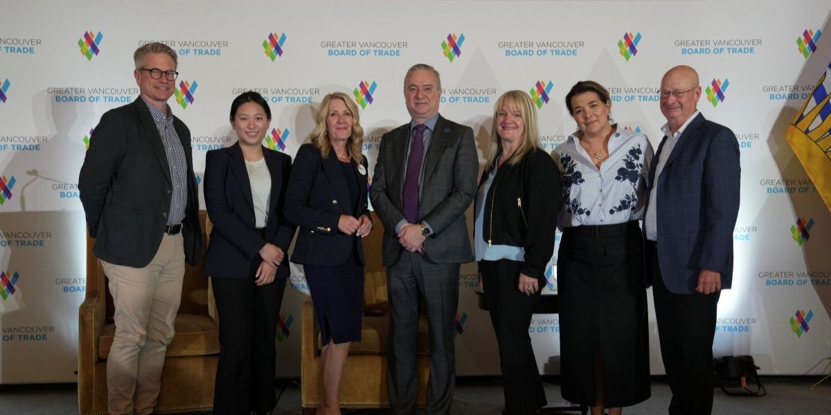 A group of seven professionally dressed individuals stand in a line on a stage in front of a backdrop that reads “Greater Vancouver Board of Trade.” The individuals, consisting of four women and three men, are smiling and posing for the photo. The backdrop features the organization's colorful logo repeated in a pattern. Two empty armchairs and part of a provincial flag are visible on either side of the group. The setting appears to be a formal event or panel discussion.