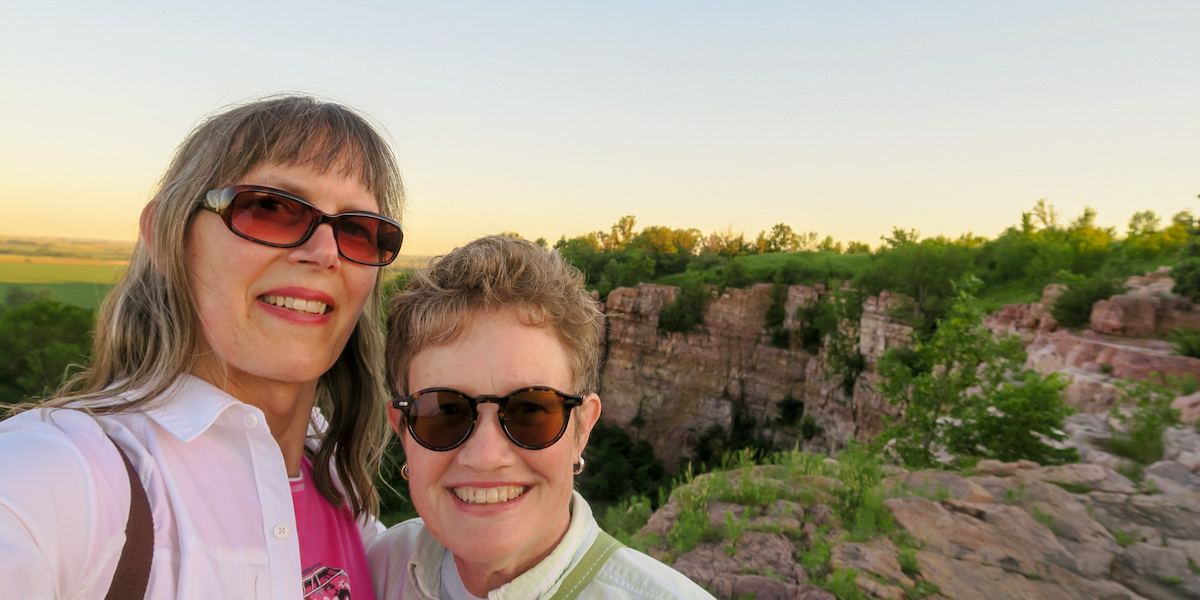 Two people smiling and taking a selfie together on a rocky hillside, surrounded by natural scenery.