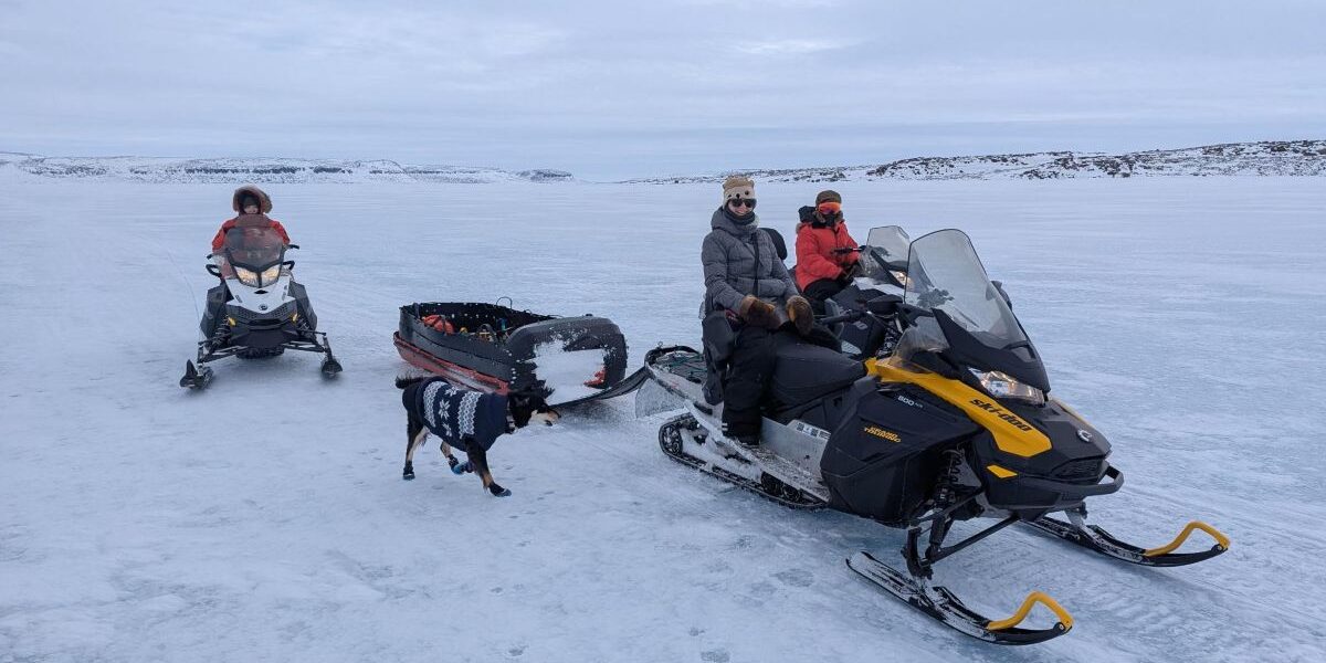 three people and a dog on snow with snowmobiles