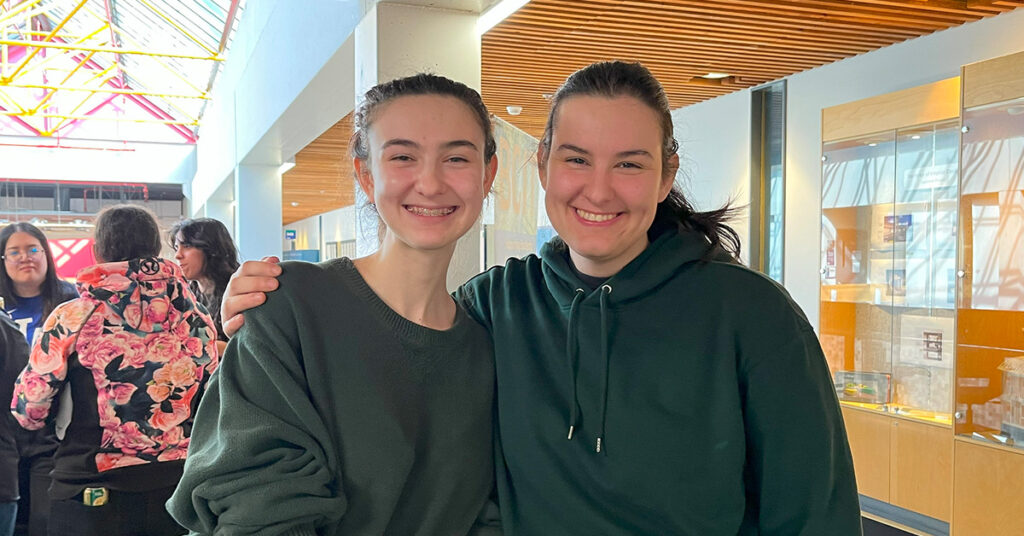 Two young women in green sweatshirts with hair in ponytails smile at the camera. The woman on the right is Tia, the subject of this story. The woman on the left is Tia's sister.