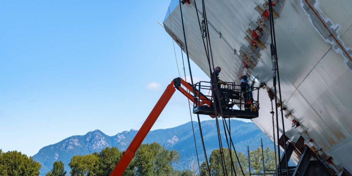 Workers welding on a large ship hull with mountains in the background.