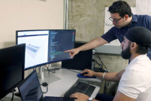 2 people in a lab one sitting and one leaning over their shoulder pointing at the computer screen