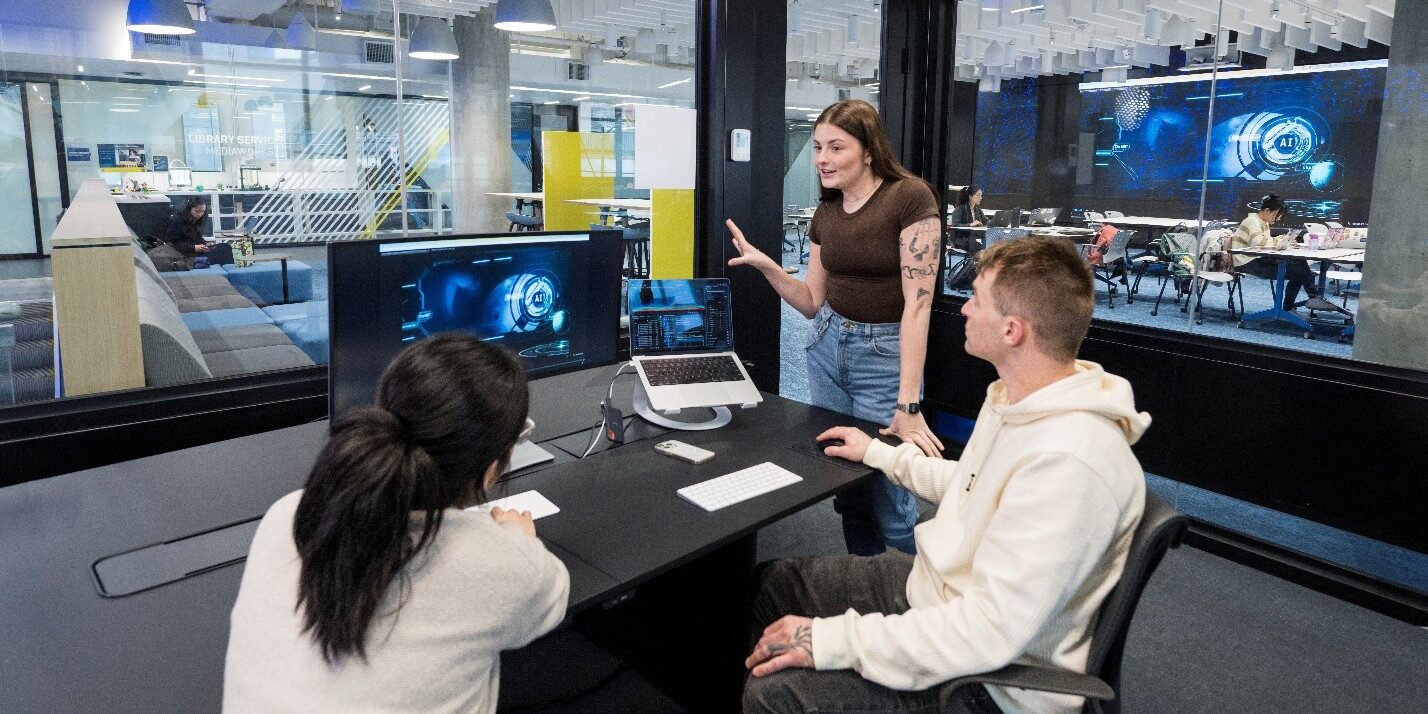 Three students around a table in a discussion. They are looking at several large and small monitors.