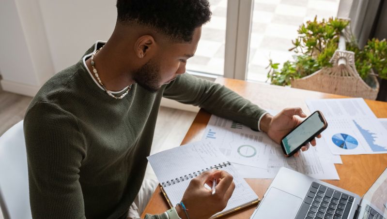 An image of a person with a phone in their hand sitting at a table leaning over a book, a laptop and some other papers. 