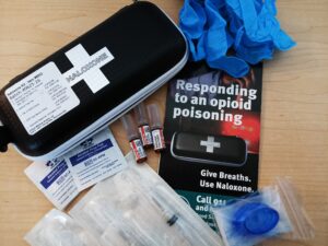 The components of a naloxone kit, including 3 needles, a pamphlet that says "Responding to opioid poisoning" and gloves.