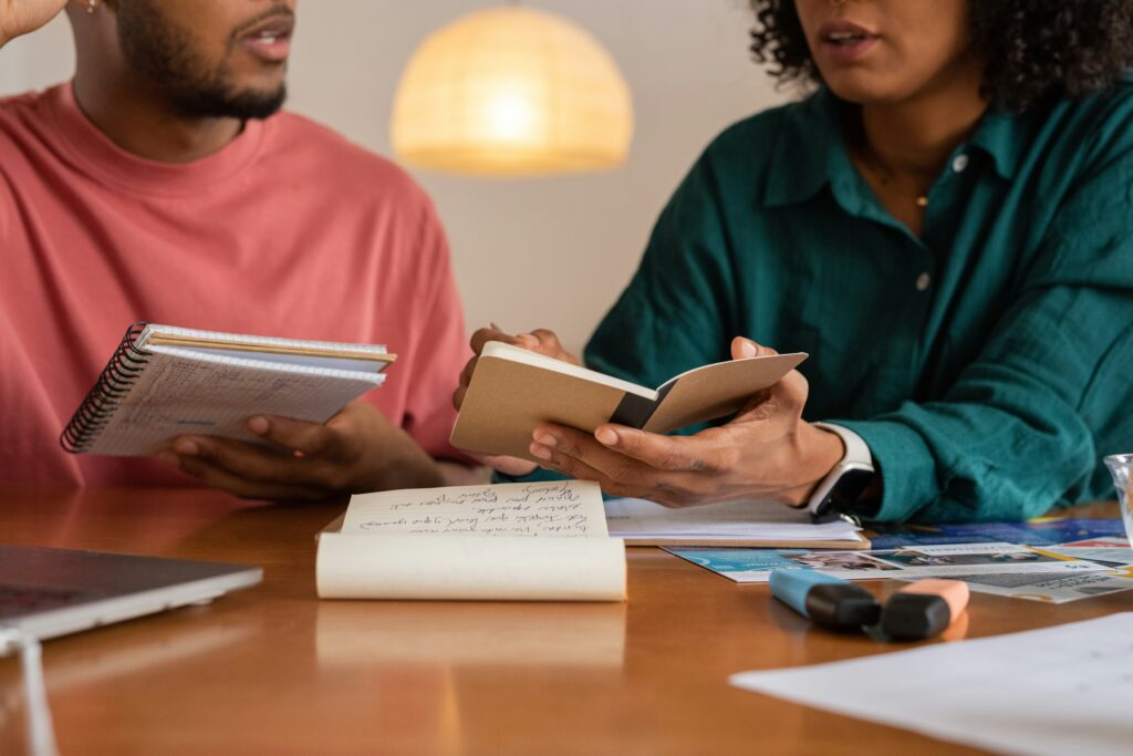 Two people sitting at a table talking with books and notebooks on the table.