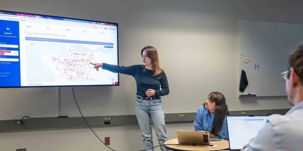female student gestures towards a projected map of Vancouver showing data and red location dots, as another computing student and instructor look on