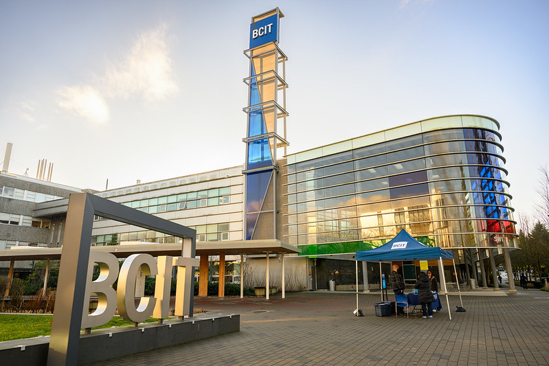 An image of the BCIT sign with SW01 building in the background. In the foreground is a booth with people around it.