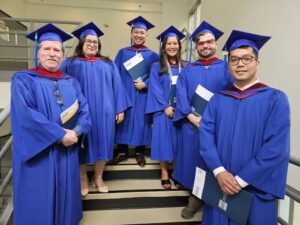 Six TMGT BTech graduates stand together smiling on a beige stairway, in formal blue cap and gown while holding their credentials at the BCIT Convocation Ceremony June 2026.