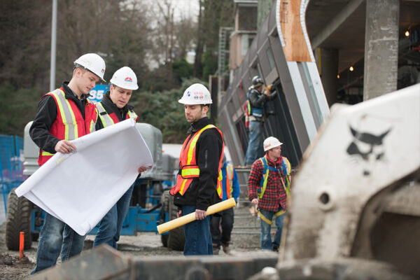 Construction workers in hard hats and high-visibility vests reviewing blueprints at an active building site.