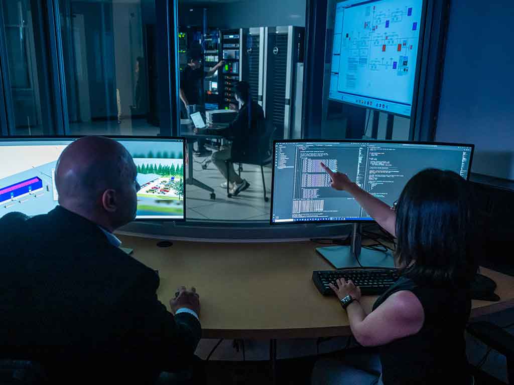 Two researchers review digital twin software on large computer monitors in a control room overlooking a server and equipment lab. One researcher points to code on the screen while another observes, with network infrastructure and a digital system diagram visible in the background.