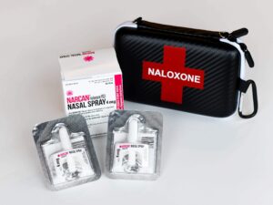 2 packages of nasal naloxone are in front of a box that says "NARCAN nasal spray", next to a black case that says "naloxone" in a red cross.