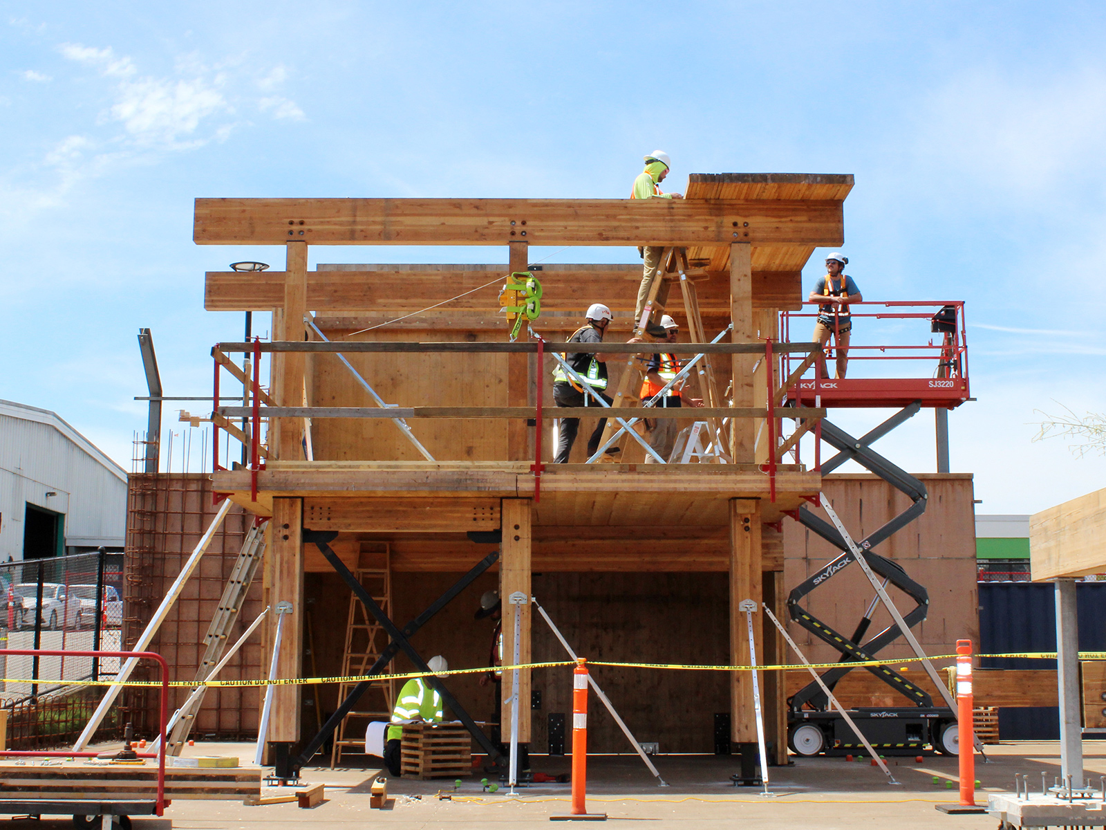 Participants working on a two-storey mass timber structure at BCIT campus, with a scissor lift and scaffolding visible alongside the glulam frame.
