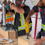 Participants drilling into a mass timber beam using a portable drill press and handheld drill, with wood shavings on the ground.