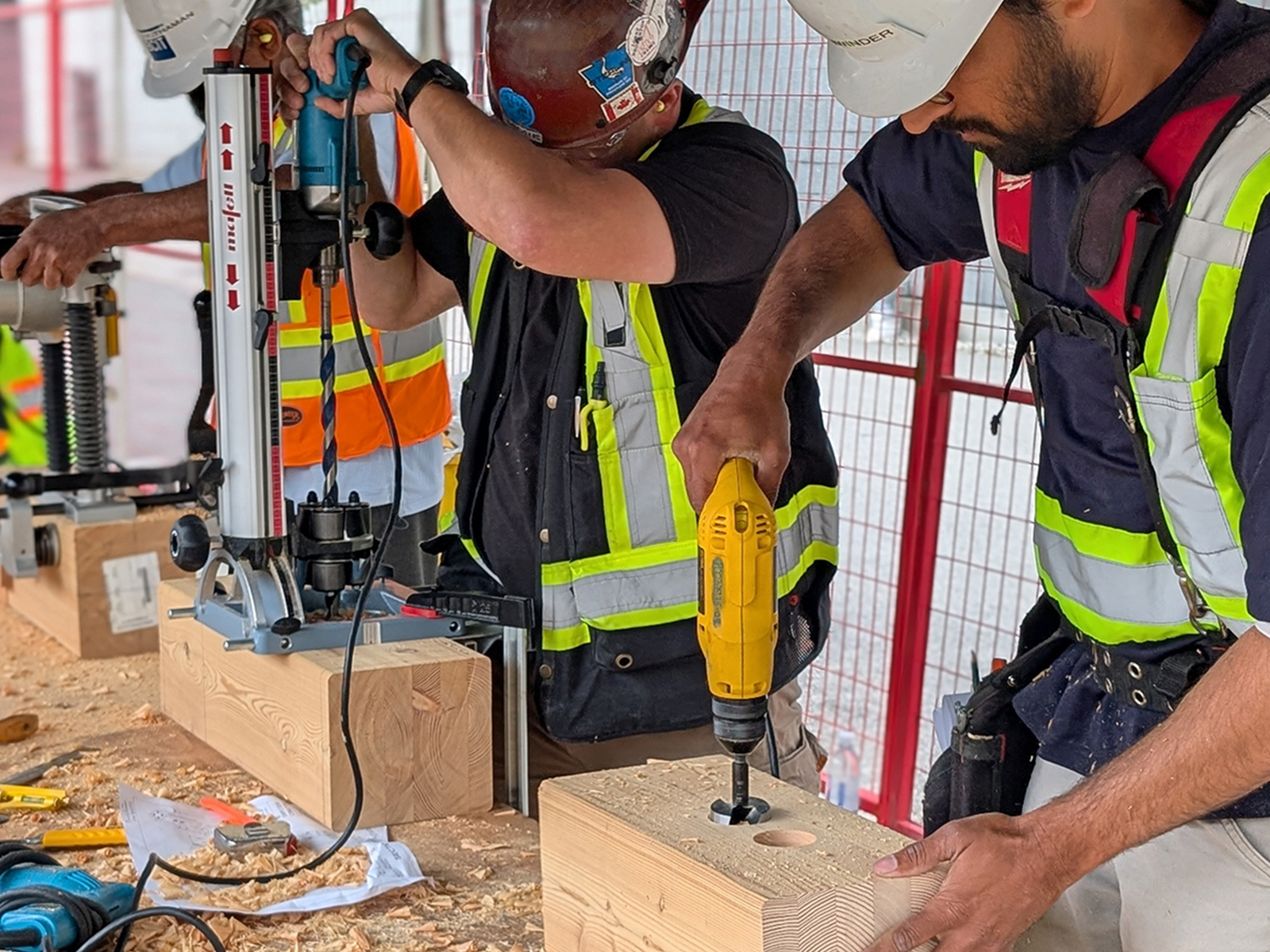 Participants drilling into a mass timber beam using a portable drill press and handheld drill, with wood shavings on the ground.
