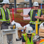 An instructor in a BCIT hard hat explaining a detail to a group of participants gathered around a concrete base plate on site.