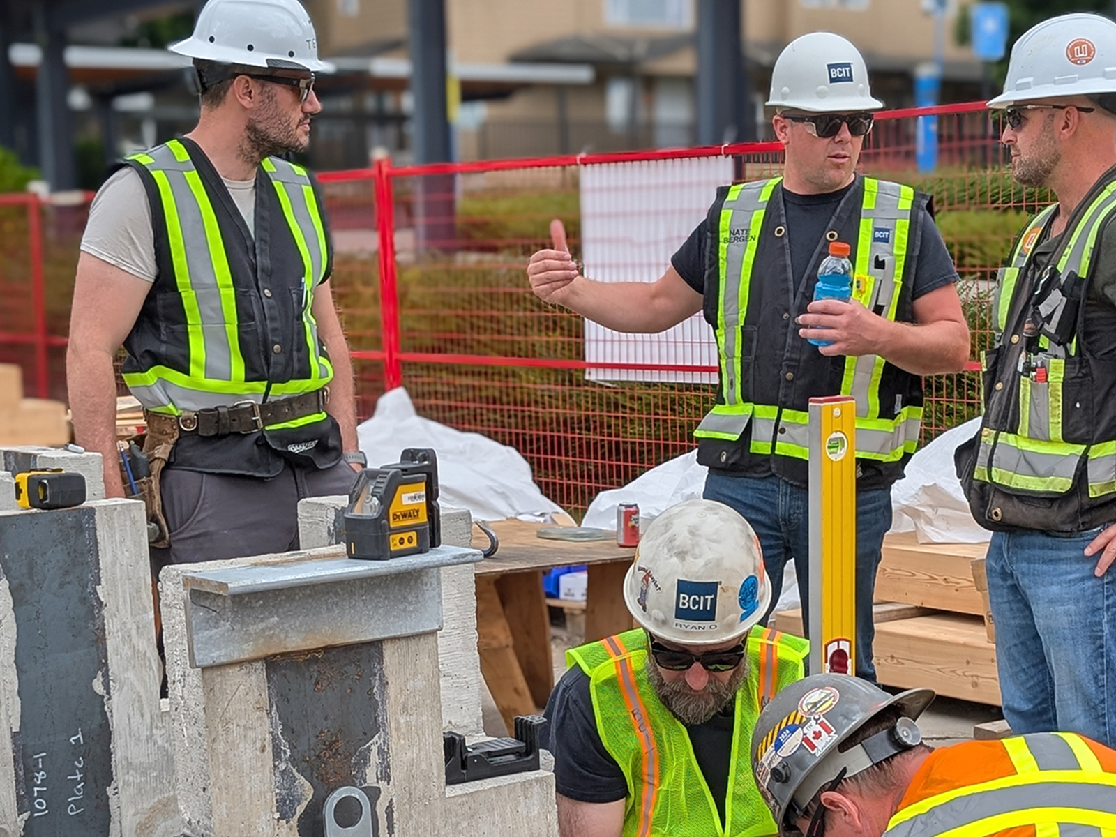 An instructor in a BCIT hard hat explaining a detail to a group of participants gathered around a concrete base plate on site.
