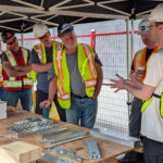 An instructor presenting mass timber connection hardware to a group of participants gathered around a workbench under a canopy.