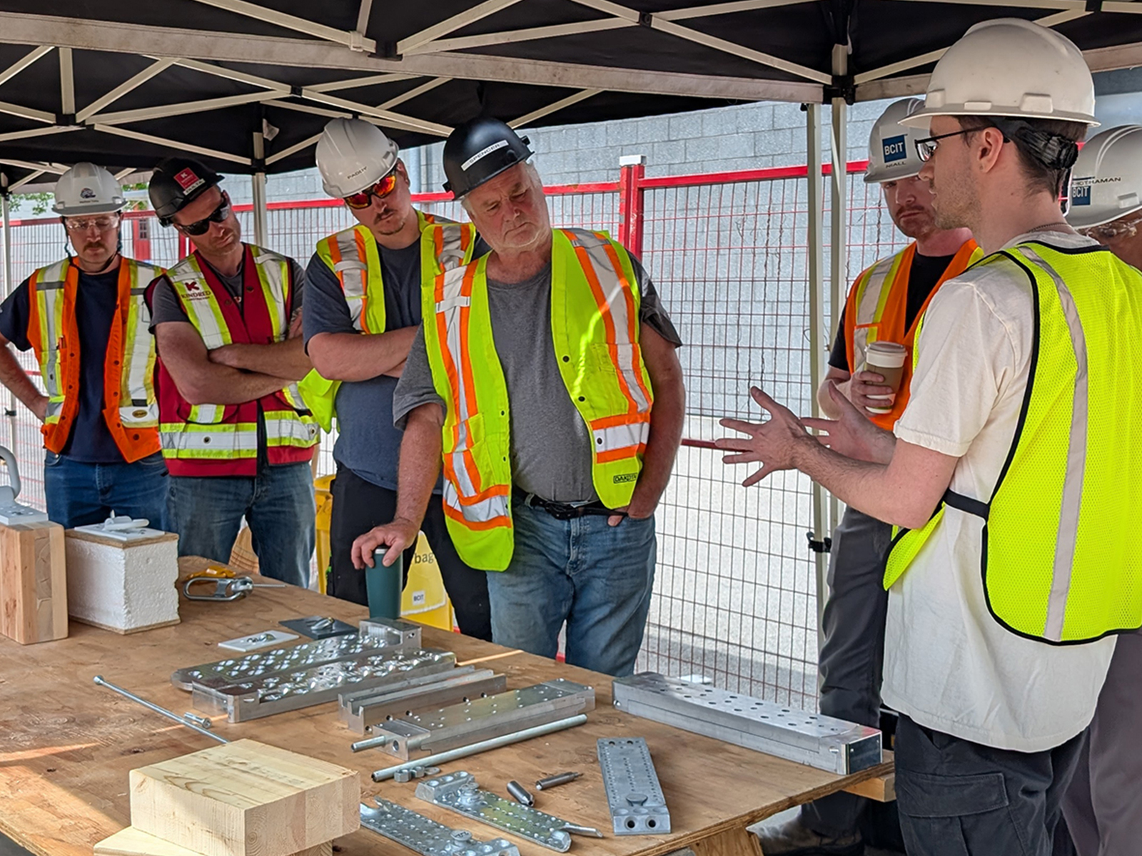 An instructor presenting mass timber connection hardware to a group of participants gathered around a workbench under a canopy.