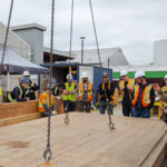 Participants gathered around a large CLT panel suspended by chains from a crane, receiving a briefing on rigging and lifting procedures.