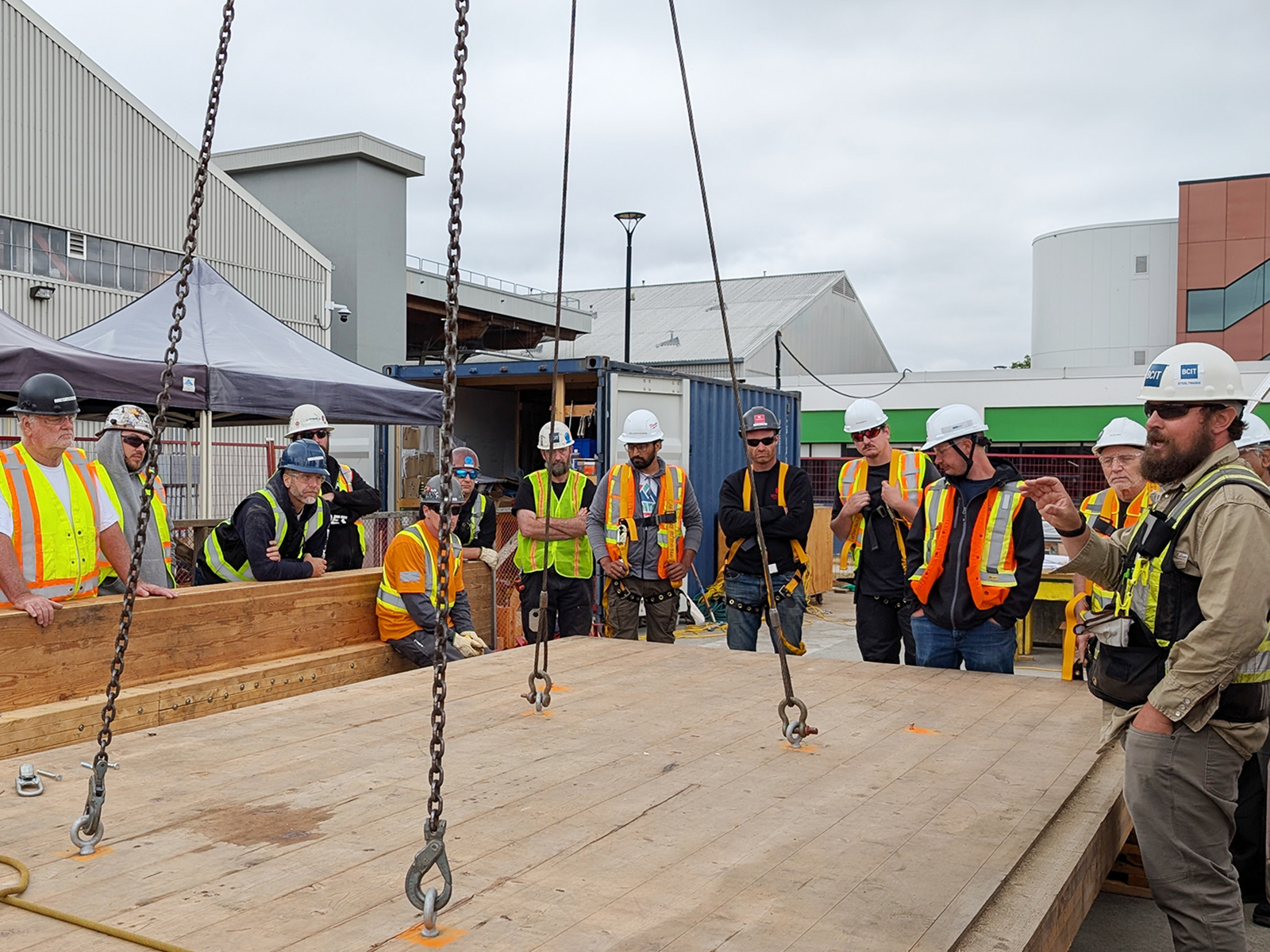 Participants gathered around a large CLT panel suspended by chains from a crane, receiving a briefing on rigging and lifting procedures.