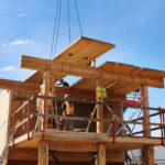 A CLT panel being lifted by crane chains onto the mass timber structure, with two participants guiding it into position from the second level.