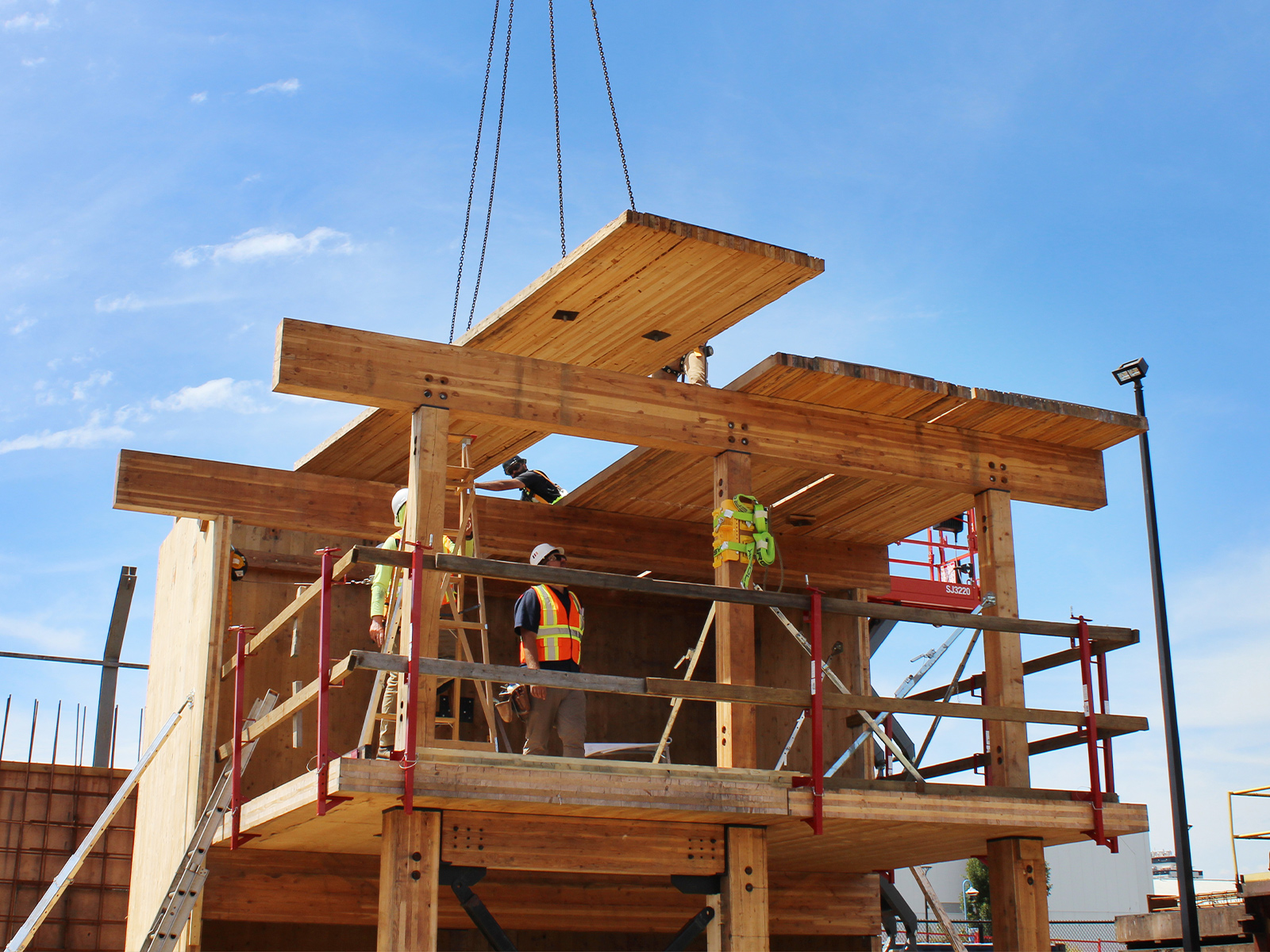 A CLT panel being lifted by crane chains onto the mass timber structure, with two participants guiding it into position from the second level.