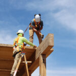 Two participants in safety gear working at the top of a glulam beam, one hammering a connection while the other steadies the structure from a ladder.