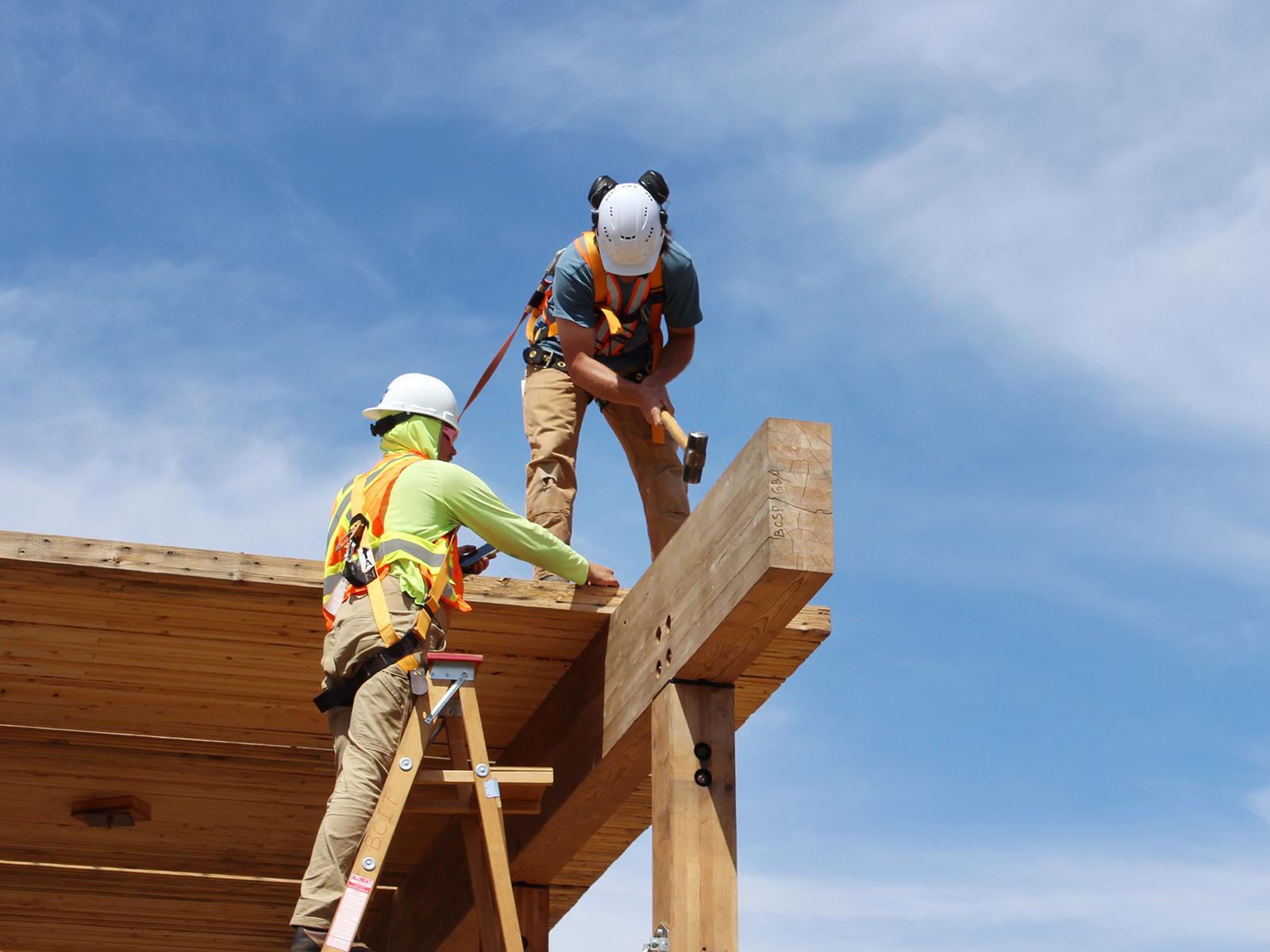 Two participants in safety gear working at the top of a glulam beam, one hammering a connection while the other steadies the structure from a ladder.