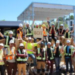 Group photo of practicum participants celebrating in front of the completed two-storey mass timber structure, holding a BCIT sign.