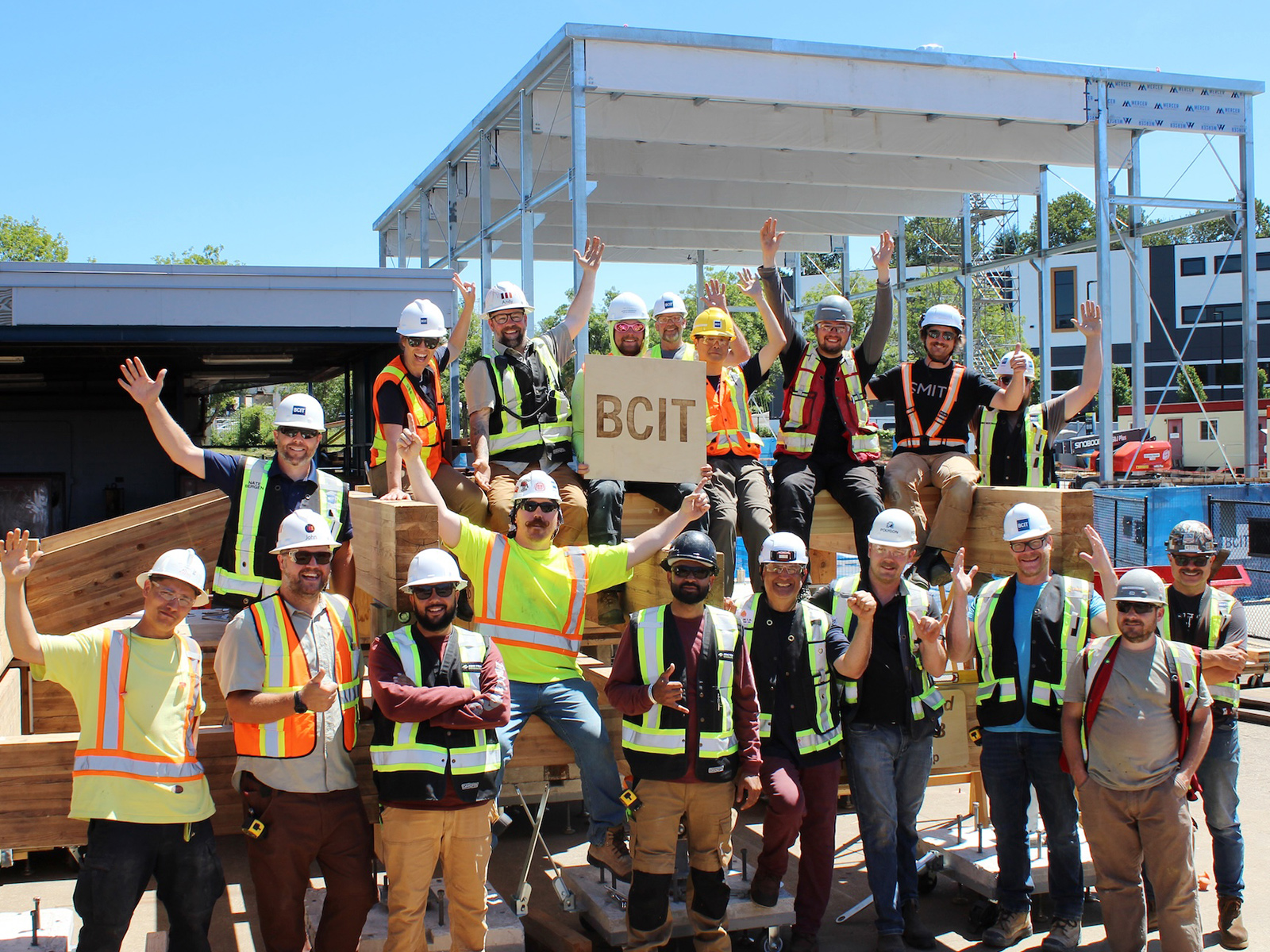 Group photo of practicum participants celebrating in front of the completed two-storey mass timber structure, holding a BCIT sign.