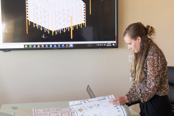 A woman with long hair reviews architectural floor plans on a table while a 3D structural timber frame model is displayed on a wall-mounted screen behind her.